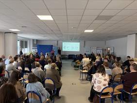 Pokaż zdjęcie: Sala konferencyjna z liczną publicznością siedzącą na krzesłach ustawionych w rzędach. Na scenie prelegentka prowadzi prezentację przy ekranie z wyświetlonym slajdem, obok stoją roll-upy organizatorów i partnerów wydarzenia.