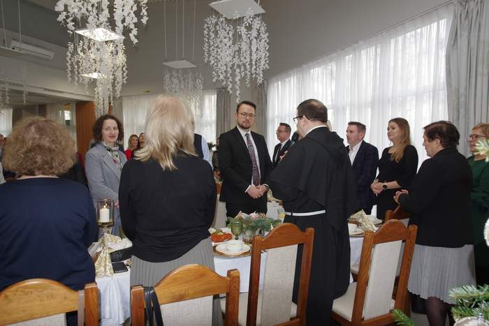 Pokaż zdjęcie: Uczestnicy spotkania wigilijnego stoją przy uroczyście nakrytym stole w jasnej sali. W centrum uwagi znajduje się mężczyzna w czarnym habicie zakonnym, który przemawia do zebranych, trzymając mikrofon. Pozostali goście, ubrani w stroje wizytowe, słuchają go z uwagą. Nad stołem wiszą ozdobne, białe żyrandole przypominające zwisające kwiaty lub płatki śniegu, a na stole widoczne są stroiki bożonarodzeniowe.