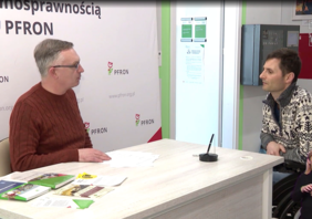 Pokaż zdjęcie: Dwóch mężczyzn siedzi przy białym biurku podczas rozmowy. Po lewej stronie mężczyzna w okularach i rdzawym swetrze, po prawej mężczyzna w jasnym swetrze we wzory, siedzący na wózku inwalidzkim. W tle znajduje się ścianka reklamowa z logo PFRON (Państwowy Fundusz Rehabilitacji Osób Niepełnosprawnych) oraz adresem strony internetowej. W prawym dolnym rogu widoczna jest mała sylwetka tłumaczki języka migowego.