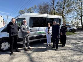Pokaż zdjęcie: Przed samochodem stoi kilka osób. Wszyscy są elegancko ubrani. Na pojeździe jest naklejka z logo PFRON. 