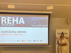 Pokaż zdjęcie: Slajd konferencji REHA for the Blind in Poland w Gdańsku 13 sierpnia 2025 z napisem Marcelina Hernik, Ekspertka ds. dostępności. Obok ekranu kobieta przy mikrofonie, w tle roll-up Fundacji Szansa dla Niewidomych