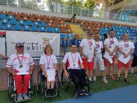 Pokaż zdjęcie: Na wózkach siedzą dwaj mężczyźni i jedna kobieta z medalami na szyi. Kobieta trzyma w ręku dyplom. Dalej w rzędzie stoją czterej mężczyźni z medalami na szyi. W tle stadion