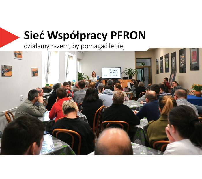 Pokaż zdjęcie: Sala wypełniona dorosłymi uczestnikami siedzącymi przy stołach, zwróconymi w stronę prowadzącego. Prelegent stoi z przodu z mikrofonem w dłoni, obok ekranu z wyświetloną prezentacją. W tle widać rośliny doniczkowe, drzwi oraz ściany ozdobione oprawionymi fotografiami. W lewym górnym rogu widnieje hasło „Sieć Współpracy PFRON – działamy razem, by pomagać lepiej”.