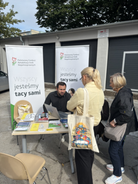 Pokaż zdjęcie: Przy stole znajdują się materiały informacyjne, ulotki i gadżety. Kilka osób rozmawia z przedstawicielami stoiska. Widoczne są roll-upy PFRON z hasłem „Wszyscy jesteśmy tacy sami”. Na pierwszym planie kobieta trzyma papierową torbę z kolorową grafiką.