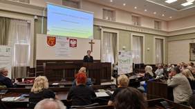 Pokaż zdjęcie: kobieta stoi na mównicy, przed nią słuchacze zasiadający na sali obrad w Urzędzie Miasta w Kielcach