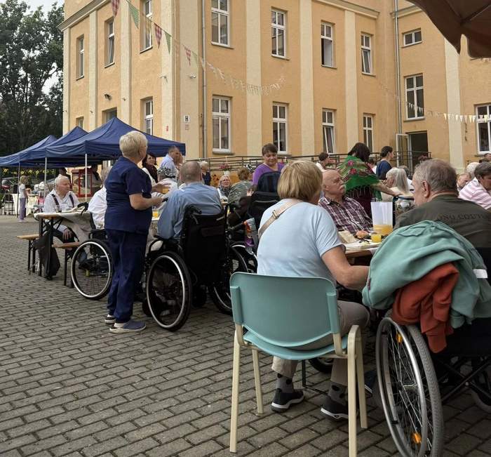 Pokaż zdjęcie: Osoby widoczne tyłem siedzące przy stolikach, częściowo osoby na wózkach. Wydarzenie odbywa się na zewnątrz budynku, na podwórku Domu.