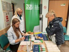 Pokaż zdjęcie: W budynku ZUS-u w Wieruszowie trwają Dni Seniora. Przy stole znajdują się trzy kobiety: jedna z nich siedzi na krześle, a dwie stoją. Panie rozmawiają ze sobą. 