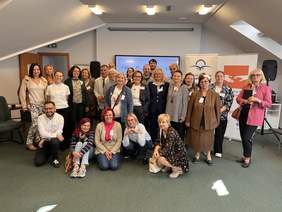 Pokaż zdjęcie: Grupa około 25 osób w sali konferencyjnej pozuje do wspólnego zdjęcia; część siedzi z przodu, większość stoi z tyłu. W tle ekran i roll-upy z logotypami organizacji, uczestnicy mają identyfikatory i uśmiechają się.