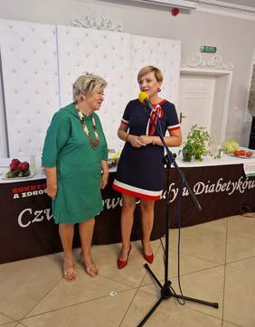 Pokaż zdjęcie: Dwie kobiety stoją przy stole z warzywami podczas wydarzenia „Czwartkowe Obiady u Diabetyków”. Jedna z nich przemawia do mikrofonu, a druga uśmiecha się w jej stronę.