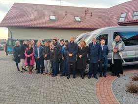Pokaż zdjęcie: Przed budynkiem są zaparkowane dwa samochody. Obok nich stoi kilkanaście osób. Wszyscy są elegancko ubrani. 