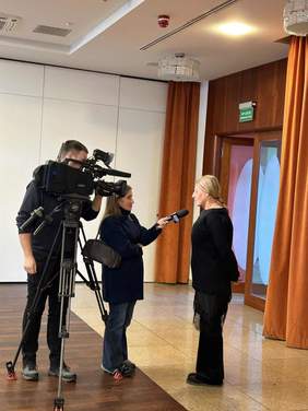 Pokaż zdjęcie: Dziennikarka nagrywa wywiad z kobietą, która jest terapeutką zajęciową. Operator kamery nagrywa rozmowę. 