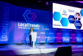 Pokaż zdjęcie: Prelegentka przemawia na scenie podczas konferencji LocalTrends – Samorządowy Kongres Finansowy. Stoi przy mównicy, za nią duży ekran z logo wydarzenia i prezentacją dotyczącą Sopotu.
