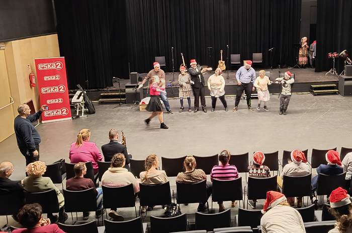Pokaż zdjęcie: Na zdjęciu widać scenę w przestrzeni teatralnej lub koncertowej. Na scenie znajduje się grupa osób, które uczestniczą w występie lub aktywności artystycznej. Wszyscy mają na głowach czerwone czapki Mikołaja, co sugeruje, że wydarzenie ma świąteczny charakter. W tle widać instrumenty muzyczne, takie jak gitary, oraz mikrofony przygotowane na statywach.  Przed sceną siedzi publiczność w rzędach krzeseł, składająca się z osób w różnym wieku. Niektórzy widzowie również noszą mikołajkowe czapki. Po lewej stronie widoczny jest baner z logo radia „Eska”. Atmosfera na zdjęciu wydaje się radosna i swobodna, a całość sprawia wrażenie świątecznego spotkania lub wydarzenia kulturalnego.