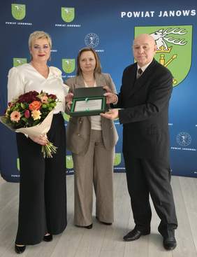 Pokaż zdjęcie: Trzy osoby, dwie kobiety i mężczyzna. Kobieta ma ręku kwiaty, druga trzyma w ręku otwarty grawerton.