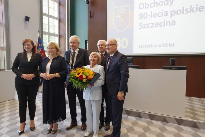 Pokaż zdjęcie: Pamiątkowe zdjęcie grupowe po wręczeniu tytułu Honorowego Ambasadora Szczecina. Barbara Jaskierska stoi w środku, trzymając bukiet kolorowych kwiatów. Po bokach stoją przedstawiciele władz miasta i organizatorzy wydarzenia. W tle widać flagi oraz ekran z 