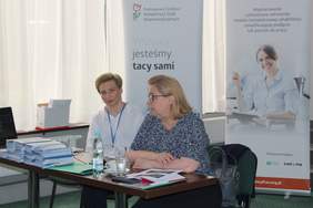 Pokaż zdjęcie: Dwie kobiety siedzą przy stoliku na którym leżą ulotki. Za nimi stoją dwa rollapy z logo Kompleksowej Rehabilitacji oraz PFRON 