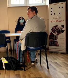 Pokaż zdjęcie: ężczyzna i kobieta siedzą na przeciwko sobie przy biurku. Na podłodze leży czarny pies przewodnik rasy labrador