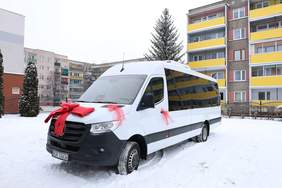 Pokaż zdjęcie: Biały bus ozdobiony czerwoną kokardą stoi na śniegu przed blokami mieszkalnymi.