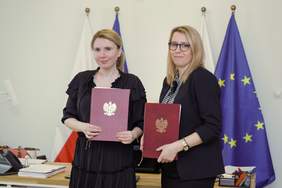 Pokaż zdjęcie: Na tle flag Polski i Unii Europejskiej stoja dwie kobiety. W rekach trzymają teczki z nadrukowanym godłem Polski.