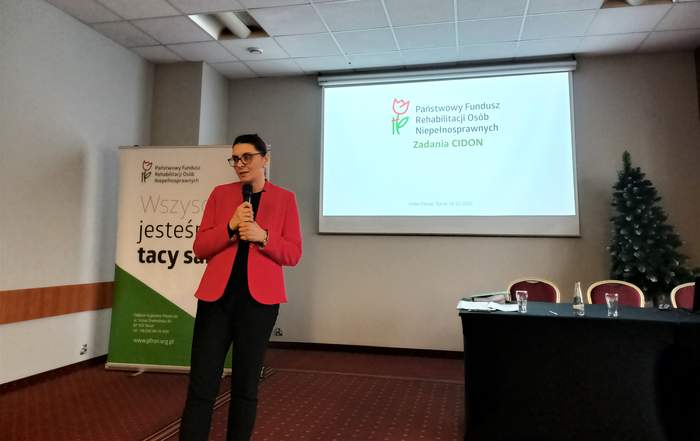 Pracownik PFRON - Anna Wankiewicz-Lewandowska - stoi przed słuchaczami i opowiada o CIDON-ie. W tle widać ekran z logo i nazwą programu oraz baner PFRON-u