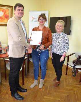 Pokaż zdjęcie: Na zdjęciu 2 kobiety i mężczyzna. Jedna z kobiet trzyma teczkę z logo PFRON. Wszyscy się uśmiechają
