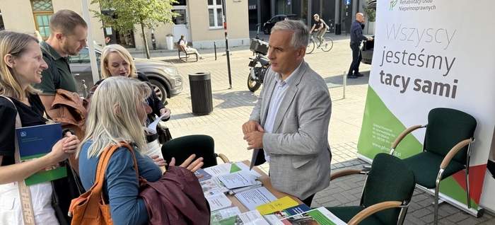 Pokaż zdjęcie: Za biurkiem stoi mężczyzna w koszuli marynarce, rozmawia ze stojącymi przed nimi ludźmi. Na stole leży dużo materiałów informacyjnych z logo PFRON. Za nim stoi duża ścianka zrobiona z 3 banerów z logo PFRON.