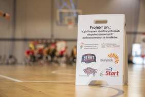 Pokaż zdjęcie: Tablica informacyjna z napisem „Projekt pn. Cykl imprez sportowych osób niepełnosprawnych” oraz logotypami partnerów, w tym Państwowego Funduszu Rehabilitacji Osób Niepełnosprawnych, Ministerstwa Sportu i Turystyki, Start Białystok, Miasta Białystok, Województwa Podlaskiego i PZSN Start. W tle rozmazany obraz zawodników na wózkach oraz kosza na hali sportowej.