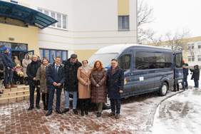 Pokaż zdjęcie: Grupa osób stoi przed budynkiem Domu Pomocy Społecznej obok nowego, przystosowanego mikrobusu; zimowa sceneria, lekko pada śnieg.