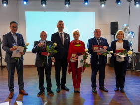 Pokaż zdjęcie: Grupa sześciu dorosłych osób stojących w rzędzie trzymających białe róże i statuetki z podziękowaniem za wspólny wkład w historię Zakładu Aktywności Zawodowej. Mężczyźni są ubrani w garnitury, a kobiety elegancko – jedna w czerwonym stroju, druga w ciemniejszym. W tle widać jasny ekran, światła sceniczne i srebrne balony. 