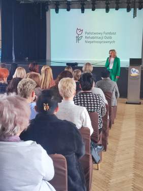 Pokaż zdjęcie: Zdjęcie sali konferencyjnej robione z tyłu sali, na przodzie prelegent kobieta w zielonym garniturze.
