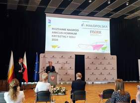 Pokaż zdjęcie: Marszałek Województwa Małopolskiego - Witold Kozłowski otwiera galę rozdania nagród. Stoi na środku sali, w tle napis Rozdanie nagród Amicus Hominum oraz Kryształy Soli 