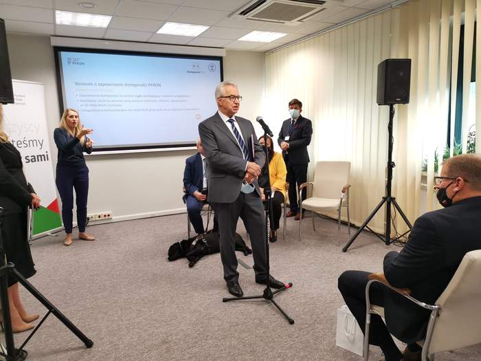 Pokaż zdjęcie: Przemawiający do mikrofonu w sali konferencyjnej mężczyzna w szarym garniturze. W tle prezentacja multimedialna, tłumaczka języka migowego, mężczyzna z psem przewodnikiem i kilka zgromadzonych osób.