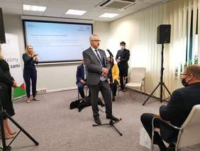 Pokaż zdjęcie: Przemawiający do mikrofonu w sali konferencyjnej mężczyzna w szarym garniturze. W tle prezentacja multimedialna, tłumaczka języka migowego, mężczyzna z psem przewodnikiem i kilka zgromadzonych osób.