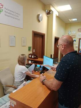 Pokaż zdjęcie: W sekretariacie Łódzkiego Oddziału PFRON znajdują się dwie osoby: kobieta i mężczyzna. Pani siedzi na krześle i patrzy w ekran monitora. Pan stoi przy ladzie i podaje jakieś dokumenty. Na biurku stoją także różne urządzenia biurowe. 