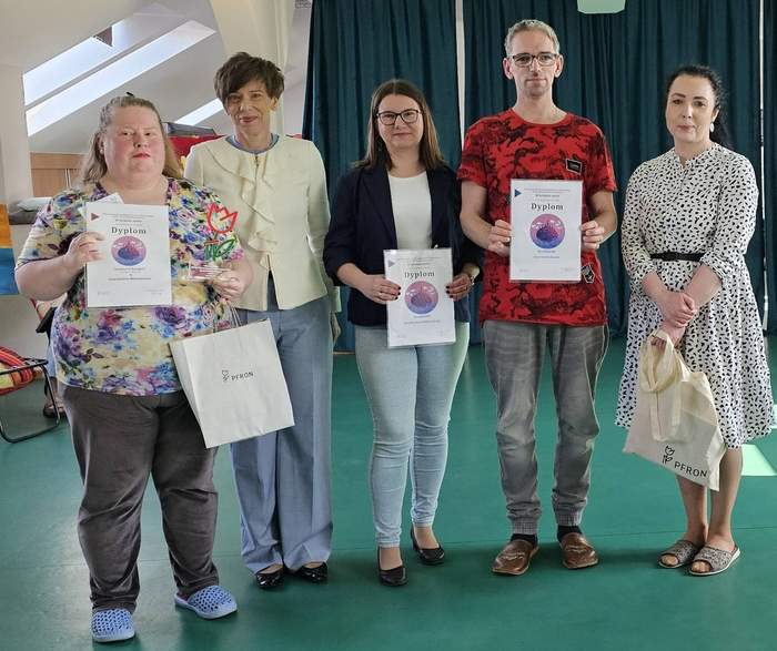 Pokaż zdjęcie: Na zdjęciu od lewej: kobieta w kwiecistej bluzce i niebieskich klapkach, trzymająca dyplom, białą torbę PFRON oraz statuetkę tulipana. Obok niej kobieta z krótkimi ciemnymi włosami w jasnej marynarce, a następnie kobieta w okularach i czarnej marynarce, trzymająca dyplom. Kolejną osobą jest mężczyzna w czerwonej koszulce z dyplomem w dłoni. Grupę zamyka kobieta w białej sukience w grochy, trzymająca białą torbę z logo PFRON.