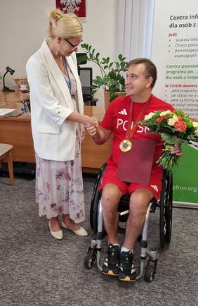 Pokaż zdjęcie: Urszula Kierznowska wręcza Rafałowi Czuperowi list gratulacyjny oraz kwiaty w siedzibie PFRON. Czuper, w sportowym stroju Polski, siedzi na wózku inwalidzkim, z medalem na szyi i bukietem kwiatów w ręku.