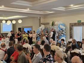 Pokaż zdjęcie: Grupa kilkudziesięciu osób znajduje się w sali restauracyjnej udekorowanej balonami. Część z tych osób stoi w pobliżu wejścia do sali, a część siedzi przy stolikach.