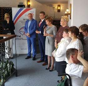 Pokaż zdjęcie: Kameralne pomieszczenie, kilkoro stojących osób, jedna na wózku inwalidzkim. 