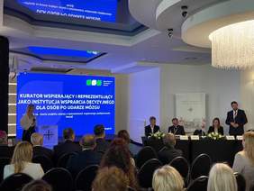Pokaż zdjęcie: Scena podczas trwania konferencji. Z lewej strony kobieta wygłasza prezentację przy mównicy, za nią na ekranie wyświetlony jest slajd zatytułowany: „Kurator wspierający i reprezentujący jako instytucja wsparcia decyzyjnego dla osób po udarze mózgu”. Po prawej stronie za stołem prezydialnym siedzi pięciu prelegentów, a jeden z mężczyzn stoi obok, przemawiając. Uczestnicy konferencji siedzą przodem do sceny. Wnętrze eleganckie, z nowoczesnym oświetleniem i żyrandolem.
