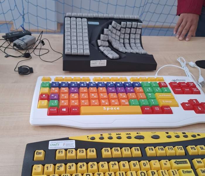 Pokaż zdjęcie: Na stole leżą trzy różne klawiatury, dostosowane do potrzeb osób z niepełnosprawnościami. Jedna jest ergonomiczna i rozdzielona, druga ma kolorowe klawisze, a trzecia duże, żółte klawisze. To przykład sprzętu wspomagającego dostępność cyfrową.