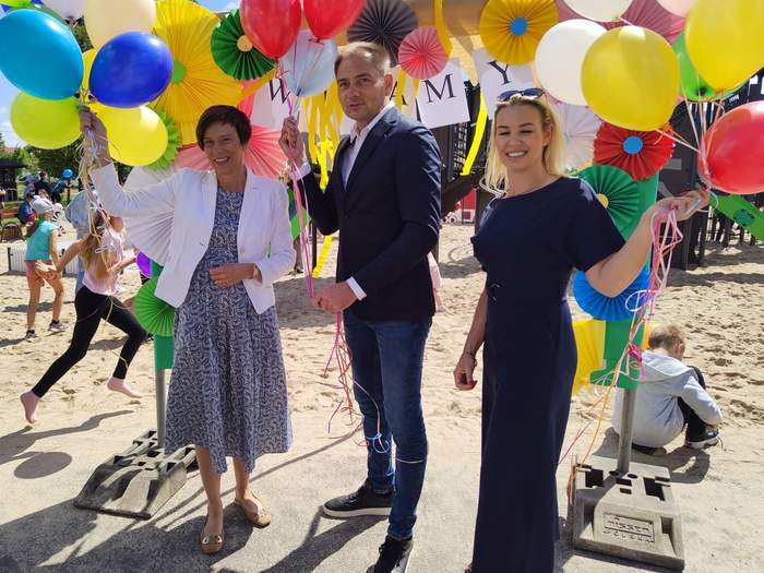 Pokaż zdjęcie: Dwie uśmiechnięte kobiety oraz jeden mężczyzna stoją przed wejściem na plac zabaw i trzymają w dłoniach kolorowe balony. Wchodząc na plac zabaw możemy dostrzec podwieszone kolorowe dekoracje z papieru, napis: „Witamy” oraz bawiące się dzieci.