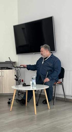 Pokaż zdjęcie: Mężczyzna siedzi na krześle i wpatruje się w ekran ustawionego przed nim laptopa. Na drugim planie szafka i telewizor wiszący na ścianie.