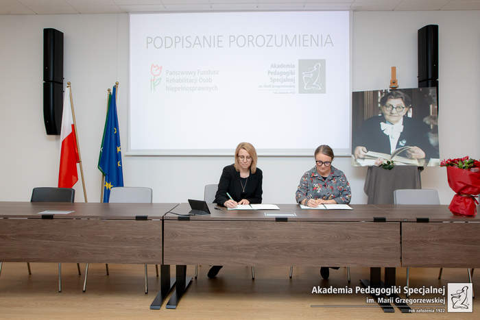 Pokaż zdjęcie: Długi stół prezydialny, przy którym siedzą dwie kobiety i podpisują dokumenty. Kobieta z lewej strony: w czarnym garniturze, blond włosy do ramion, nosi okulary. Kobieta z prawej strony w szarym żakiecie w kolorowe wzory, ciemne włosy spięte do tyłu w kok, nosi okulary. W tle z lewej strony, za stołem, stoją flagi Polski i Unii Europejskiej, z prawej strony zdjęcie Marii Grzegorzewskiej, po środku tablica z wyświetlonym slajdem. Na slajdzie napis Podpisanie Porozumienia, pod napisem logo PFRON i APS. W prawym dolnym logu zdjęcia jest logotyp APS.