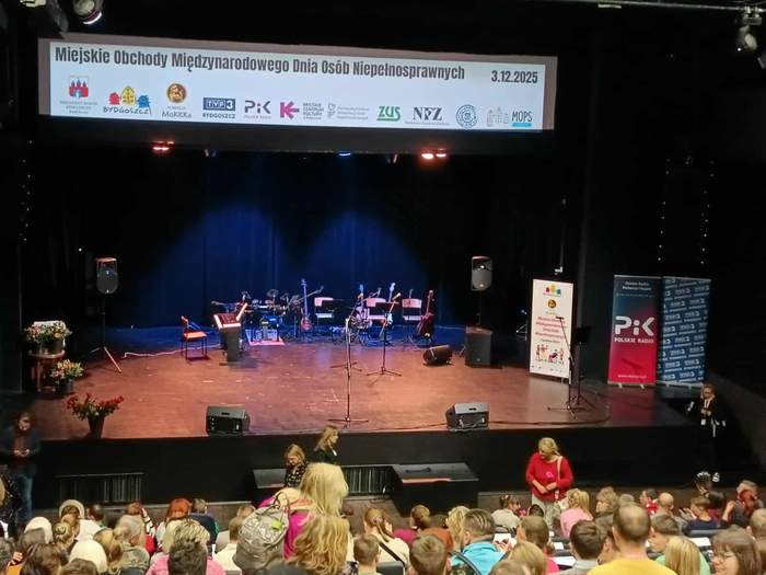 Oświetlona scena, na widowni przed sceną siedzą kobiety i mężczyźni, zdjęcie wykonane od tyłu