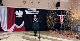 Pokaż zdjęcie: Kobieta w zielonej sukience przemawia, trzymając mikrofon. Za nią widnieje napis „XI Przegląd Pieśni Patriotycznej - Jestem Polakiem”, godło Polski oraz biało-czerwona flaga. Po prawej stronie znajdują się dekoracje z brzozowych drzew oraz krzyż ozdobiony biało-czerwoną wstążką.