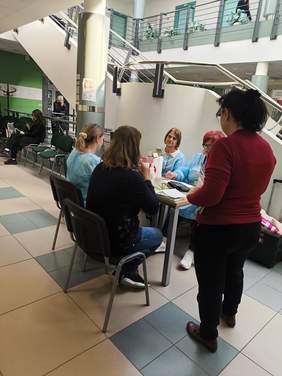 Pokaż zdjęcie: Przy biurku siedzą trzy kobiety ubrane w niebieskie fartuchy oraz jedna kobieta, której wykonywane jest badanie. Kobieta w czerwonej marynarce stoi przy biurku.