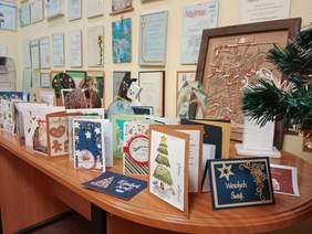 Pokaż zdjęcie: Na stoliku leży mnóstwo kartek z życzeniami świątecznymi i noworocznymi. Mają różne wielkości i są bardzo barwne. Na ścianie nad stołem wiszą dyplomy i gratulacje. 