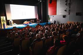 Pokaż zdjęcie: Szeroki plan widowni teatru lub sali kinowej, wypełnionej ludźmi. Na scenie po lewej stronie widać dekoracje imitujące pejzaż wodny i ruiny oraz postacie w strojach scenicznych. Na dużym ekranie w tle wyświetlony jest napis w języku polskim: "MIĘDZYNARODOWY DZIEŃ OSÓB Z NIEPEŁNOSPRAWNOŚCIAMI". Po prawej stronie sceny stoi mężczyzna w garniturze.