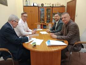 Pokaż zdjęcie: Przy podłużnym stole siedzi czterech mężczyzn. Podpisują umowę. W tle widać regał, na którym znajdują się różne odznaki, statuetki, medale, ramki z dyplomami. 