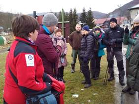 Pokaż zdjęcie: Grupa osób stojących na dworze ubranych stosownie do niskiej temperatury. Niektóre osoby mają kijki trekkingowe. 